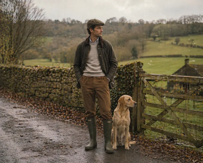 British countryside: waxed olive Barbour jacket, tattersall shirt, lambswool jumper, moleskin trousers, green Hunter wellies, tweed flat cap, Labrador at side.