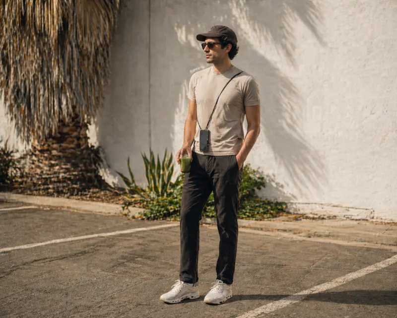 LA wellness casual: sand performance tee, black Lululemon ABC trousers, tonal On Cloud trainers, cap, crossbody phone lanyard, matcha in hand, palm shadows on stucco.
