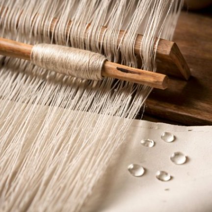Macro of a wooden hand-loom in progress — vertical warp threads in cream linen with a wooden shuttle.