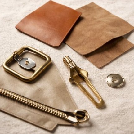 A still-life of non-textile clothing materials on cream linen — leather and suede swatches, brass buckle and clasp, snap fastener, zip.